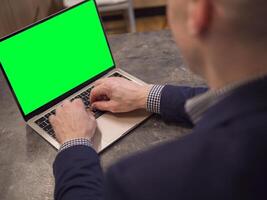 Businessman sitting at table working on laptop with vibrant green screen, typing on keyboard. Over-the-shoulder view shows focus on laptop tasks, Concept modern technology Remote work. photo