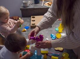 .Mother and her two children enjoy quality time together, engaging in creative play with colorful modeling clay at their kitchen table. Modeling clay and fun tools Hand Motor Concept photo