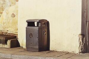 Black plastic garbage can with lid standing near the wall of a light yellow building in evening sunlight photo