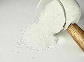 Pile of salt food flavoring ingredient spilled from glassware cup with wooden handle isolated on horizontal white background. photo