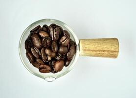 Pile of brown dry coffee beans in glass container saucer with wooden handle isolated on horizontal white background from above top angle view. photo