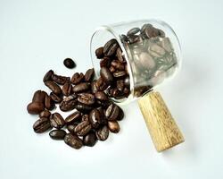 Bunch of spilled dark brown coffee beans from small glassware container with wooden handle isolated on horizontal white background. photo