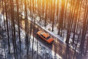 A vibrant orange car drives through a snow-covered forest, illuminated by golden sunlight, creating a warm winter ambiance photo