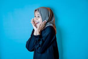 Portrait of Asian woman wearing a hijab, striking a surprised pose with both hands near her cheeks. Isolated against a blue background. photo
