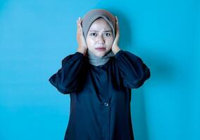 Portrait of a happy Asian woman wearing a hijab, posing because of the noise and covering her ears with both hands. Isolated against a blue background. photo