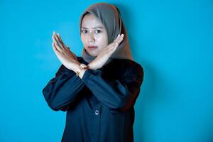 A woman in a hijab and black outfit crosses her arms in an X gesture against a blue background, expressing a sign of refusal or disagreement. photo