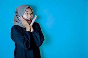 Portrait of Asian woman wearing a hijab, striking a surprised pose with both hands near her cheeks. Isolated against a blue background. photo