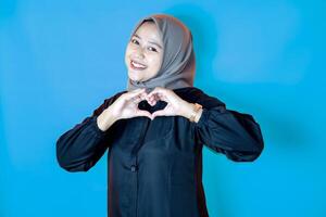 Portrait of a happy Asian woman wearing a hijab, posing with both hands forming a heart shape in front of her chest. Isolated against an blue background. photo