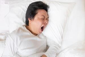 An Asian man is captured in a moment of genuine yawn as he rests on a soft white bed and pillow. The image evokes a sense of contagious yawning, inviting viewers to join in the action. photo