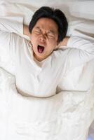 An Asian man is captured in a moment of genuine yawn as he rests on a soft white bed and pillow. The image evokes a sense of contagious yawning, inviting viewers to join in the action. photo
