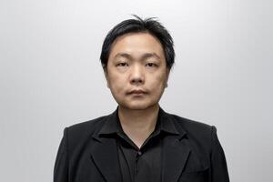 Asian man in his middle age, dressed in a stylish black suit and shirt, poses against a neutral white-gray backdrop. photo