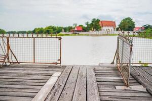 The old wooden dock with decayed iron fences beside a river in Thailand. A temple at the serene riverside. photo