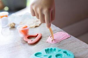 A kid playing and holding a plastic yellow knife with the right hand to cut and create a shape on the pink play dough. A green mold and a pink heart shaped mold was placed on a wooden table. photo