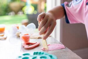 A kid playing and holding a plastic yellow knife with the right hand to cut and create a shape on the pink play dough. A green mold and a pink heart shaped mold was placed on a wooden table. photo