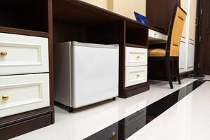 mini fridge in low angle view, inside it is empty shelves placed under wooden counter and next to wood closet and chair in a luxury hotel room. photo