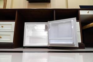 mini fridge in low angle view, inside it is empty shelves placed under wooden counter and next to wood closet and chair in a luxury hotel room. photo