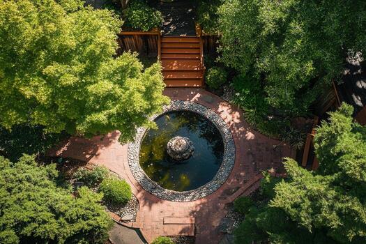 Aerial view of a tranquil courtyard garden with a circular fountain, surrounded by lush greenery and a wooden staircase. photo