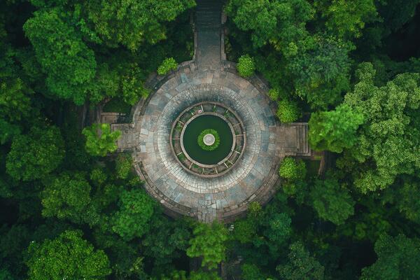 Aerial view of a circular stone structure in a lush green forest, featuring a small pond at its center.