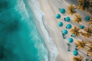 Aerial view of turquoise ocean waves lapping a white sand beach lined with palm trees and blue umbrellas. photo