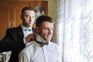Two men are getting dressed up, one of them is wearing a bow tie photo