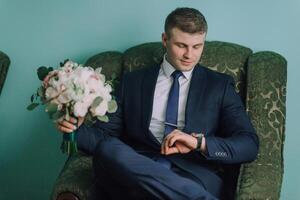 A man in a suit and tie is sitting in a green chair and looking at his watch photo