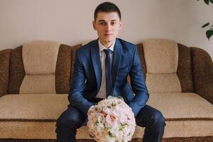 A man in a suit and tie sits on a couch holding a bouquet of flowers photo