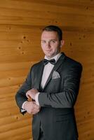 A man in a black suit and bow tie is standing in front of a wooden wall photo