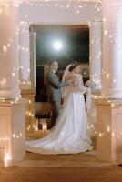 A bride and groom stand in front of a building with lights on photo