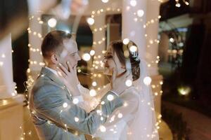 A bride and groom are embracing in front of a lighted backdrop photo
