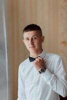 A man in a white shirt and black bow tie is standing in front of a window photo