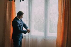 A man in a suit is standing in front of a window, looking at his watch photo