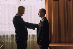 Two men are standing in a room, one of them is wearing a suit photo