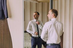 A man is getting ready for work and is adjusting his tie in front of a mirror photo