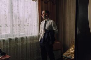 A man in a suit is standing in front of a window, looking out at the street photo