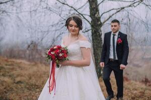 A bride and groom are standing in a field with a red bouquet and a red tie photo