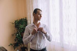 A man in a white shirt is standing in front of a window, adjusting his collar photo