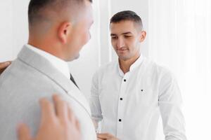 Two men are standing in front of a mirror, one of them is wearing a suit photo