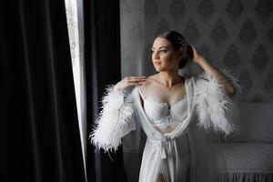 A woman in a white gown with feathers on it stands in front of a window photo