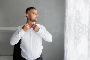 A man in a white shirt is getting dressed and looking out the window photo
