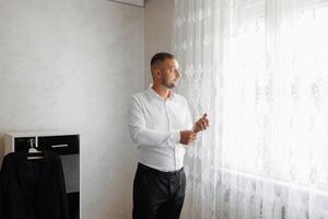 A man in a white shirt is standing in front of a window, adjusting his tie photo