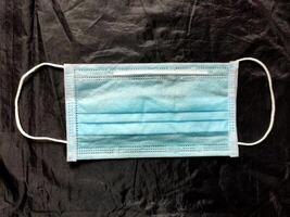 A blue disposable surgical mask with elastic ear loops and a pleated design is set against a dark background, symbolizing health, hygiene, and protection against airborne particles and droplets. photo