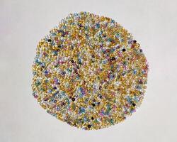 A vibrant mix of colorful seed beads in gold, silver, blue, and pink forms a circular pattern on a white background. The tiny, rounded beads with iridescent surfaces highlight crafting and creativity. photo