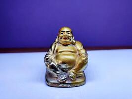 A golden Laughing Buddha figurine sits on a white surface with a purple backdrop, showcasing its joyful pose, smiling face, and plump figure, symbolizing happiness, luck, and prosperity. photo