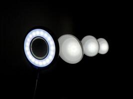 An illuminated circular LED ring light with evenly spaced bulbs shines against a dark background, accompanied by three smaller circular lights, offering a versatile setup for diffused lighting. photo