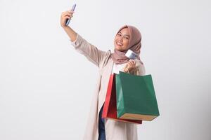 Portrait of excited Asian hijab woman in casual suit holding credit card and shopping bags while taking selfie with mobile phone. Shopaholic girl concept. Isolated image on white background photo