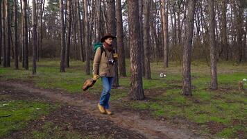 A backpacker with a guitar and a camera walks along a trail in the forest. Travel concept. Slow motion video