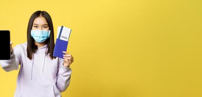 Happy korean girl in face mask, showing smartphone screen, app interface and passport with tickets for vacation, booking trip, yellow background photo