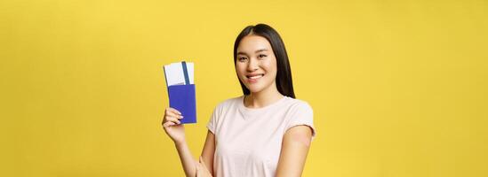 Smiling asian woman showing passport with tickets, arm with patch after covid 19 vaccination, vaccinating campaign for tourists, yellow background photo