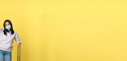 Full length shot of enthusiastic korean girl enjoying vacation, posing with suitcase, wearing face medical mask, travelling abroad during covid pandemic, yellow background photo