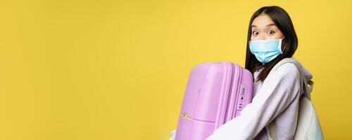 Surprised asian girl tourist, wear medical face mask, looking amazed and impressed at camera, carry suitcase in hands, yellow background photo